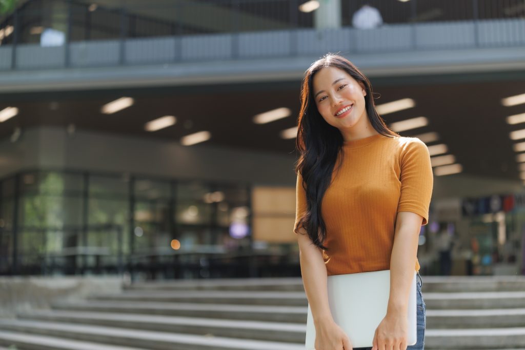 Smiling university student holding laptop is standing in front of university building
